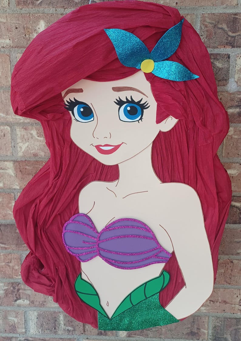 Puede incluir: Una pi&ntilde;ata con forma de sirena de dibujos animados con cabello largo y voluminoso de papel crep&eacute; rojo, un accesorio para el cabello de color verde azulado brillante, un top de concha morado brillante y una cola verde brillante.