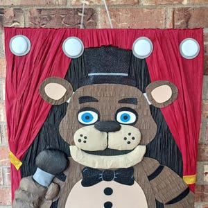 May include: A brown bear piñata with a black top hat, bow tie, and microphone. The bear is set against a red curtain backdrop with white and gray circular accents, perfect for a birthday party or themed event.