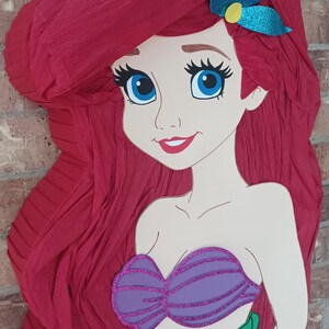 Puede incluir: Una pi&ntilde;ata con forma de sirena de dibujos animados con largo cabello de papel crep&eacute; rojo texturizado, un accesorio para el pelo azul brillante, un top de conchas morado brillante y una cola verde brillante. Tiene ojos azules.