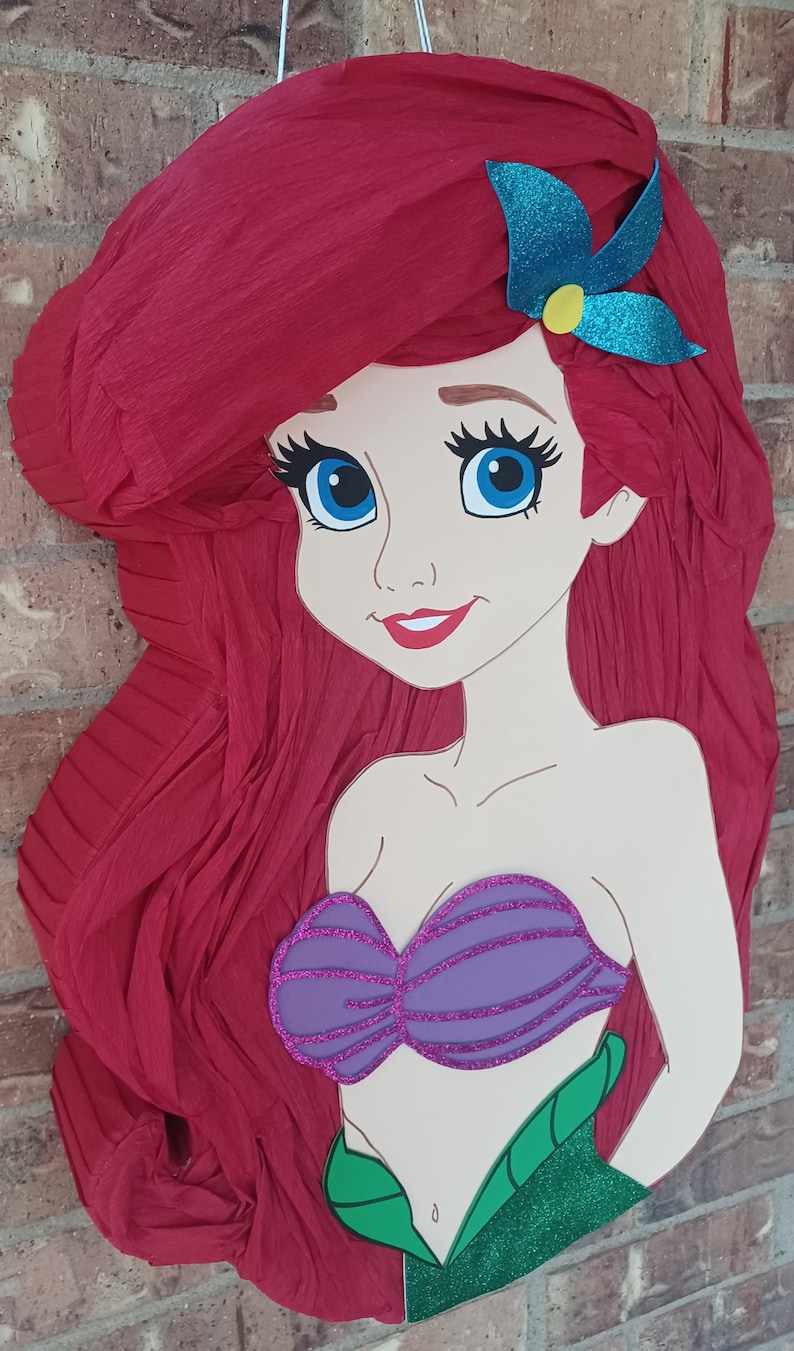 Puede incluir: Una pi&ntilde;ata con forma de sirena de dibujos animados con largo cabello de papel crep&eacute; rojo texturizado, un accesorio para el pelo azul brillante, un top de conchas morado brillante y una cola verde brillante. Tiene ojos azules.