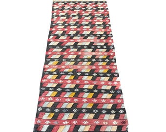 May include: A long, narrow rug with a geometric pattern in shades of red, black, white, and yellow. The rug has a fringe on both ends.