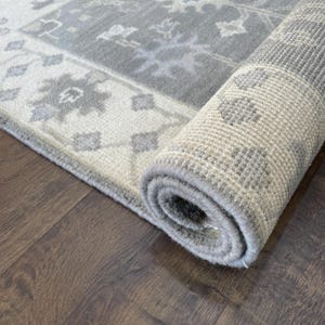 May include: A rolled up grey and white patterned rug with a subtle floral design. The rug is rolled up on a dark wood floor.
