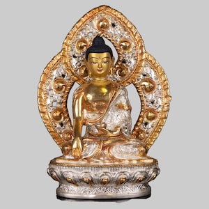 May include: A gold and silver Buddha statue sitting in a meditative pose with a decorative halo behind the head. The statue is made of metal and has a detailed design.