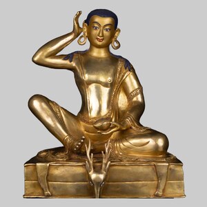 May include: A gold-colored statue of a seated figure with one arm raised to the ear. The figure has blue accents on the hair and shoulders, and wears large earrings. The statue is on a rectangular base with a deer-like figure at the front.