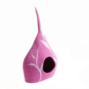 May include: A pink felt cat house, shaped like a teardrop, with a tall, pointed top. It features a round entrance and white, leaf-like patterns. The background is plain white.