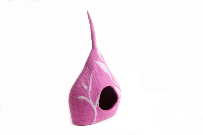 May include: A pink felt cat house, shaped like a teardrop, with a tall, pointed top. It features a round entrance and white, leaf-like patterns. The background is plain white.