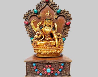 May include: A gold-colored statue of a deity sitting on a throne with a decorative base. The statue is adorned with turquoise and red gemstones. The deity has four arms and is holding a staff and a bowl.