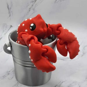 May include: A bright red crab figurine with white spots and large black eyes sits inside a small, silver metal bucket. The crab's claws are extended, and the bucket has a small handle on each side. The background is a white marble pattern.