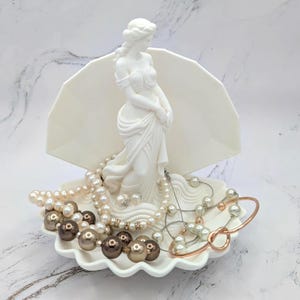 May include: A white decorative jewelry holder featuring a statue of a woman and a seashell design. The holder contains various necklaces, bracelets, and pearl beads in shades of white, brown, and gold. The jewelry holder is on a white marble surface.