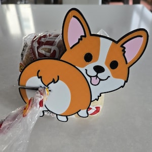 May include: A whimsical corgi-shaped bag clip, featuring orange, white, and black details. The clip is holding a clear plastic bag. The corgi has a happy expression with its tongue sticking out.