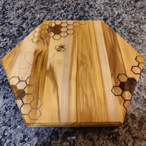 A front view of a hexagon shaped cutting board, with a honeycomb and honey bee pattern laser engraved.