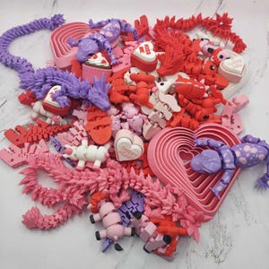 May include: A collection of Valentine's Day themed 3D printed toys and decorations. The items are in shades of pink, red, and purple and include hearts, dragons, unicorns, and a skeleton. The text "Kiss Me", "I Love You", and "Sweet Heart" is visible on some of the items.
