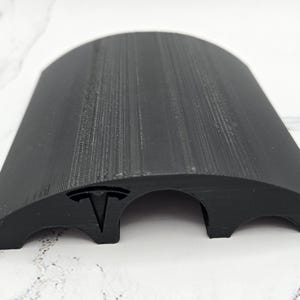 May include: Black cable management channel with a Tesla logo. The channel has two openings for cables and is designed to keep cords organized.