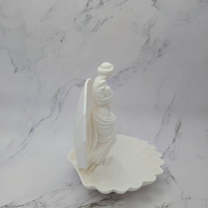 Może przedstawiać: Biała ceramiczna rzeźba kobiety stojącej w muszli. Kobieta jest odziana w powiewającą tkaninę, a muszla ma falisty brzeg. Rzeźba jest ustawiona na białym marmurowym tle.