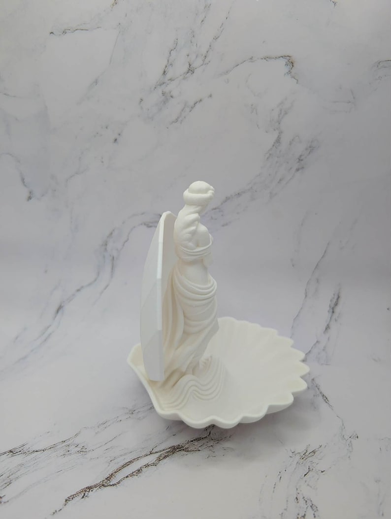 Może przedstawiać: Biała ceramiczna rzeźba kobiety stojącej w muszli. Kobieta jest odziana w powiewającą tkaninę, a muszla ma falisty brzeg. Rzeźba jest ustawiona na białym marmurowym tle.