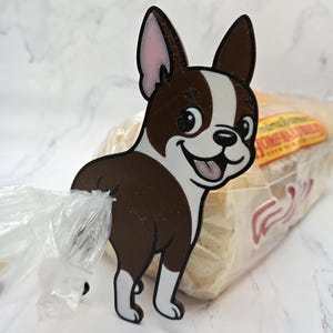 May include: A brown and white Boston Terrier dog-shaped bag clip. The clip is holding a clear plastic bag. The dog has a smiling expression and is made of wood or a similar material.