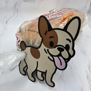 May include: A decorative bread bag clip featuring a cartoon French Bulldog design. The clip is tan, brown, and black with a pink tongue. The clip is attached to a bag of bread.