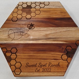 A front view of a hexagon shaped cutting board, with a honeycomb and honey bee pattern laser engraved, with a personalized name and date engraved at the bottom.