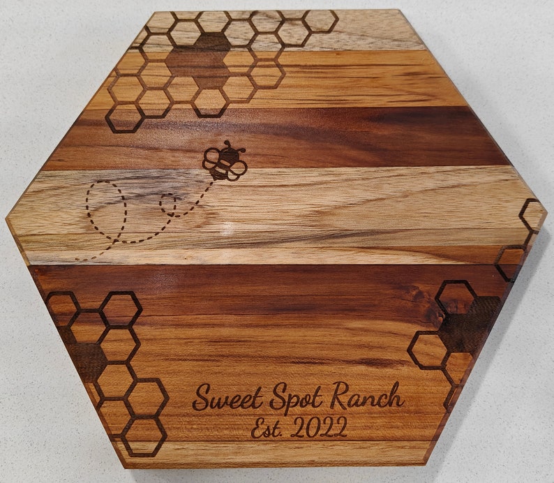 A front view of a hexagon shaped cutting board, with a honeycomb and honey bee pattern laser engraved, with a personalized name and date engraved at the bottom.