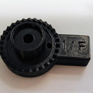 May include: Black plastic 3D printed part with a gear-like design and a rectangular section with the text "II/F" printed on it.