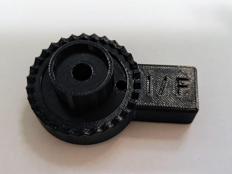 May include: Black plastic 3D printed part with a gear-like design and a rectangular section with the text "II/F" printed on it.
