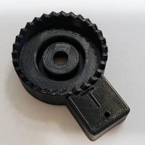 May include: A black plastic 3D printed object with a circular gear-like design and a smaller circular hole in the center. The object has a rectangular extension on the side.