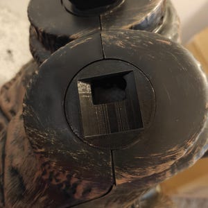 May include: Close-up of a black plastic base with a square opening. The base is attached to a larger, textured, brown object.