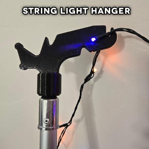 May include: A black plastic string light hanger attached to a silver telescoping pole. The hanger has a hook on the end to hold the string lights. The lights are orange and blue.
