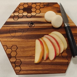 May include: A hexagonal wooden cutting board with a honey comb design and a bee illustration. The board has a knife and apple slices on it.