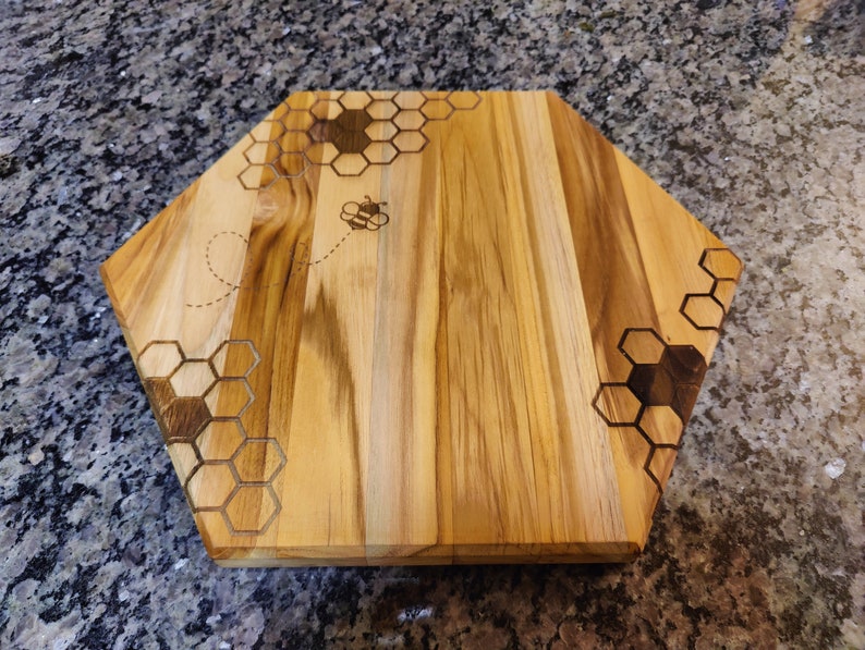 A front view of a hexagon shaped cutting board, with a honeycomb and honey bee pattern laser engraved.