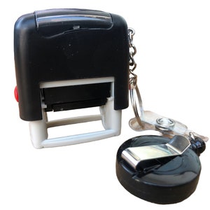 May include: A black self-inking stamp with a white frame and a red button. It is attached to a black retractable badge holder with a silver clip and a silver chain.