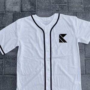 May include: White baseball jersey with black trim and a black letter K logo on the front. The jersey has a button-up front and a curved hem.
