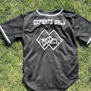 May include: Black baseball jersey with white trim and the text "EXPERTS ONLY" on the back. A white graphic of two overlapping squares with a mountain range inside is below the text.