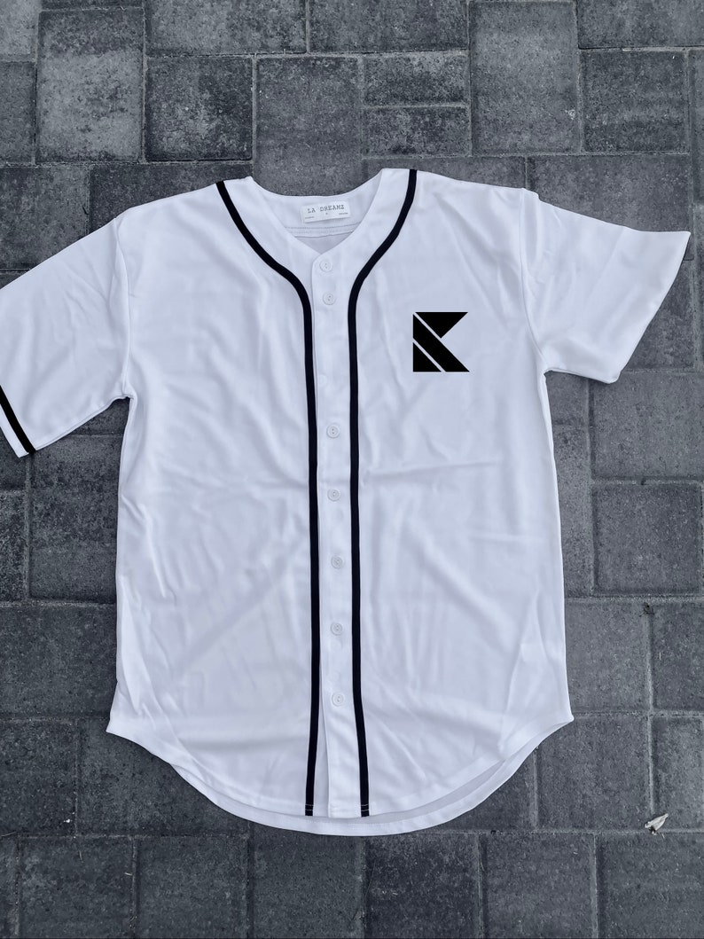 May include: White baseball jersey with black trim and a black letter K logo on the front. The jersey has a button-up front and a curved hem.