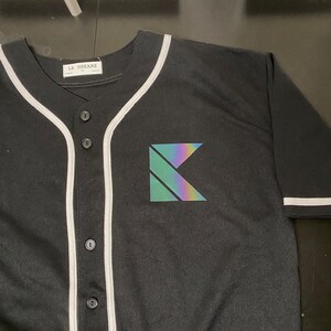 May include: Black baseball jersey with white piping and a holographic 'K' logo on the front. The jersey has a button-up front and a label that reads 'LA DREAMZ'.