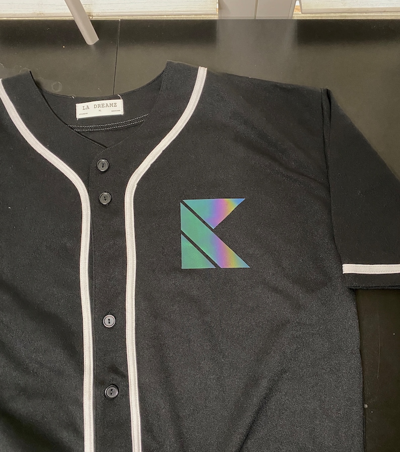May include: Black baseball jersey with white piping and a holographic 'K' logo on the front. The jersey has a button-up front and a label that reads 'LA DREAMZ'.