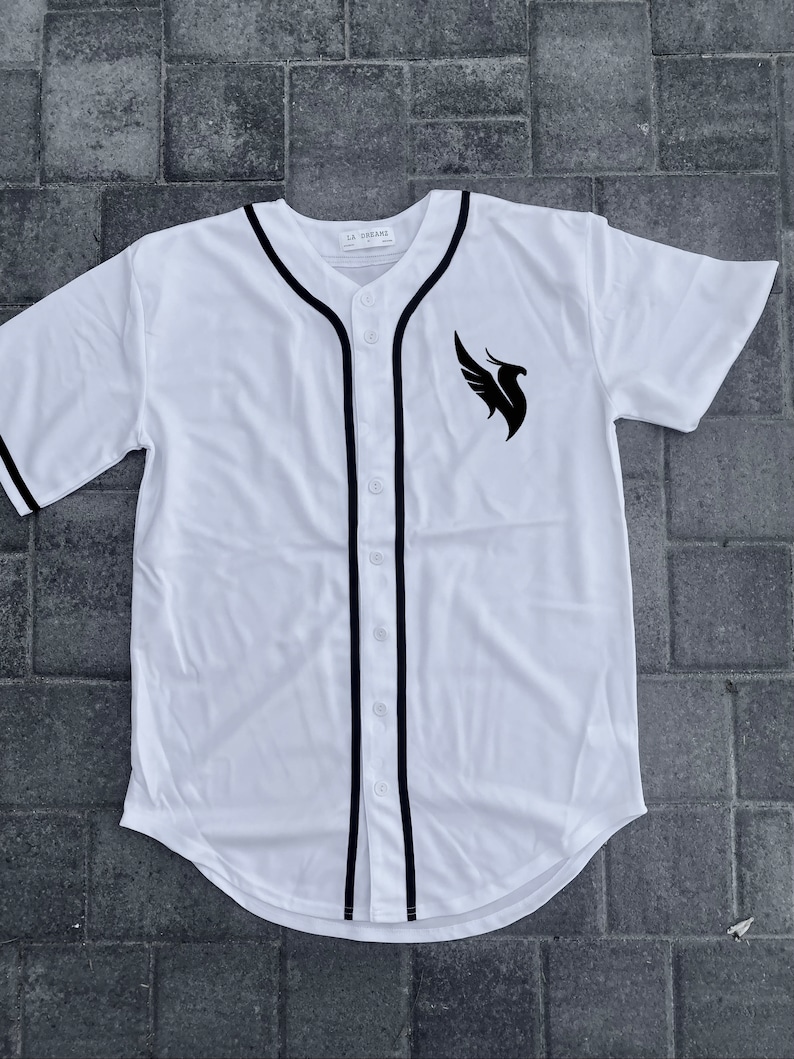 May include: White baseball jersey with black trim and a black phoenix logo on the right side. The text "LA DREAMS" is printed on the tag.