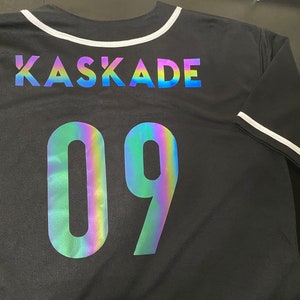 May include: Black baseball jersey with a white collar and white trim on the sleeves. The back of the jersey has the text "KASKADE" and the number "09" in iridescent rainbow lettering.