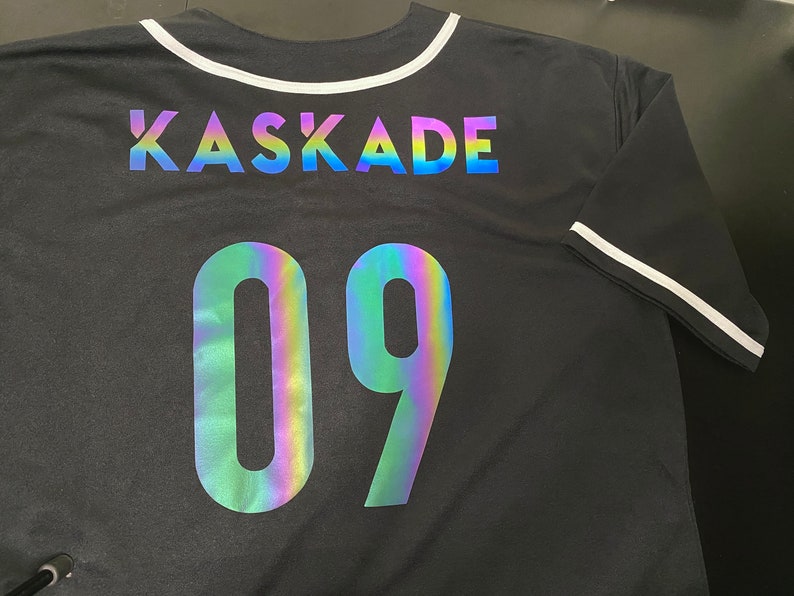 May include: Black baseball jersey with a white collar and white trim on the sleeves. The back of the jersey has the text "KASKADE" and the number "09" in iridescent rainbow lettering.
