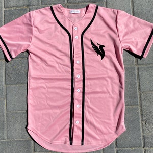 May include: A pink baseball jersey with black trim and a black phoenix logo on the front. The jersey has a button-up front and a curved hem.