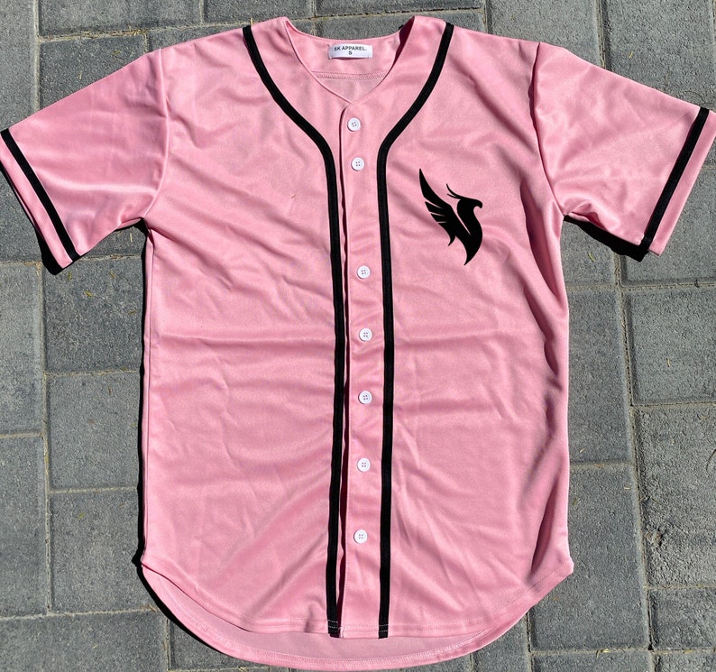 May include: A pink baseball jersey with black trim and a black phoenix logo on the front. The jersey has a button-up front and a curved hem.