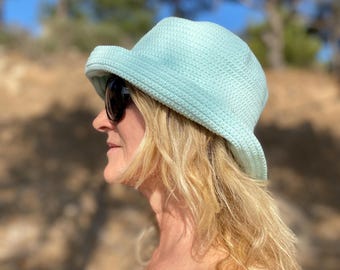 May include: A light blue crochet bucket hat with a slightly curved brim. The hat is worn by a person wearing sunglasses. The background is blurred, with trees and a blue sky visible.