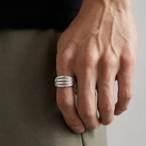 Könnte beinhalten: Ein silberner Ring mit einem strukturierten, geschichteten Design. Der Ring hat eine glatte, polierte Oberfläche und wird am Finger getragen. Das Design des Rings weist drei horizontale Bänder auf, die einen kühnen und modernen Look erzeugen. Der Ring ist ein Schmuckstück.