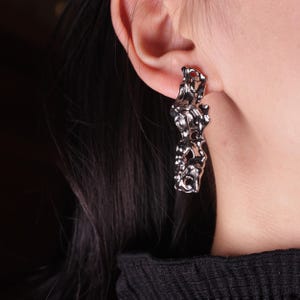 May include: A silver-coloured, abstract earring is displayed. The earring features a textured, organic design with multiple irregular shapes and a polished finish. The earring is worn on an ear, with the back of the ear and part of the person's hair visible.