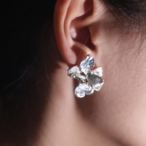 Puede incluir: Pendiente de botón plateado con forma floral. El pendiente está diseñado para parecerse a una flor con múltiples pétalos. El pendiente se lleva en la oreja, con la oreja visible en el fondo. El pendiente tiene una superficie texturizada.