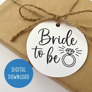 May include: A white circle tag with black text that reads "Bride to be" with a diamond ring illustration. The tag is tied with twine to a brown gift box.