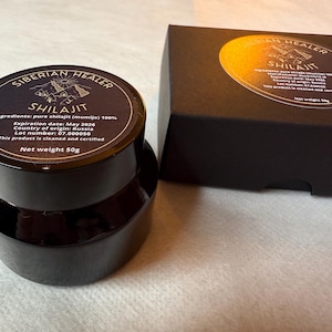 May include: A black jar with a label that reads "Siberian Healer Shilajit" with the ingredients, expiration date, country of origin, lot number, and net weight of 50g. The jar is sitting on a white surface.