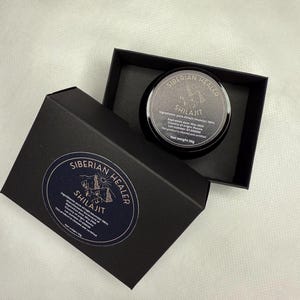 May include: A black box with a gold label that reads "SIBERIAN HEALER SHILAJIT" and contains a small black jar with a gold label that reads "SIBERIAN HEALER SHILAJIT". The jar contains a dark brown substance. The label on the jar includes the ingredients, expiration date, country of origin, lot number, and net weight.