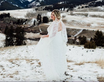 May include: A bride wearing a white lace wedding dress with long sleeves and a flowing skirt, standing in a snowy mountain landscape.