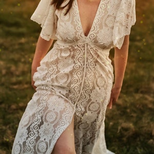 May include: A white lace maxi dress with a plunging neckline and a thigh-high slit. The dress is sheer and features a delicate floral pattern.